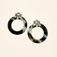 The Darling Effect's Midnight Opal Glow Getter Earrings—round, black and gray marbled with clear flower-shaped gems—are displayed on a light beige background.