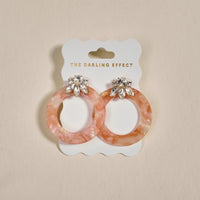 The Darling Effect’s Rose Quartz Glow Getter Earrings—pink marbled statement earrings with jeweled studs—are displayed on a white card with the brand name, set against a neutral background.