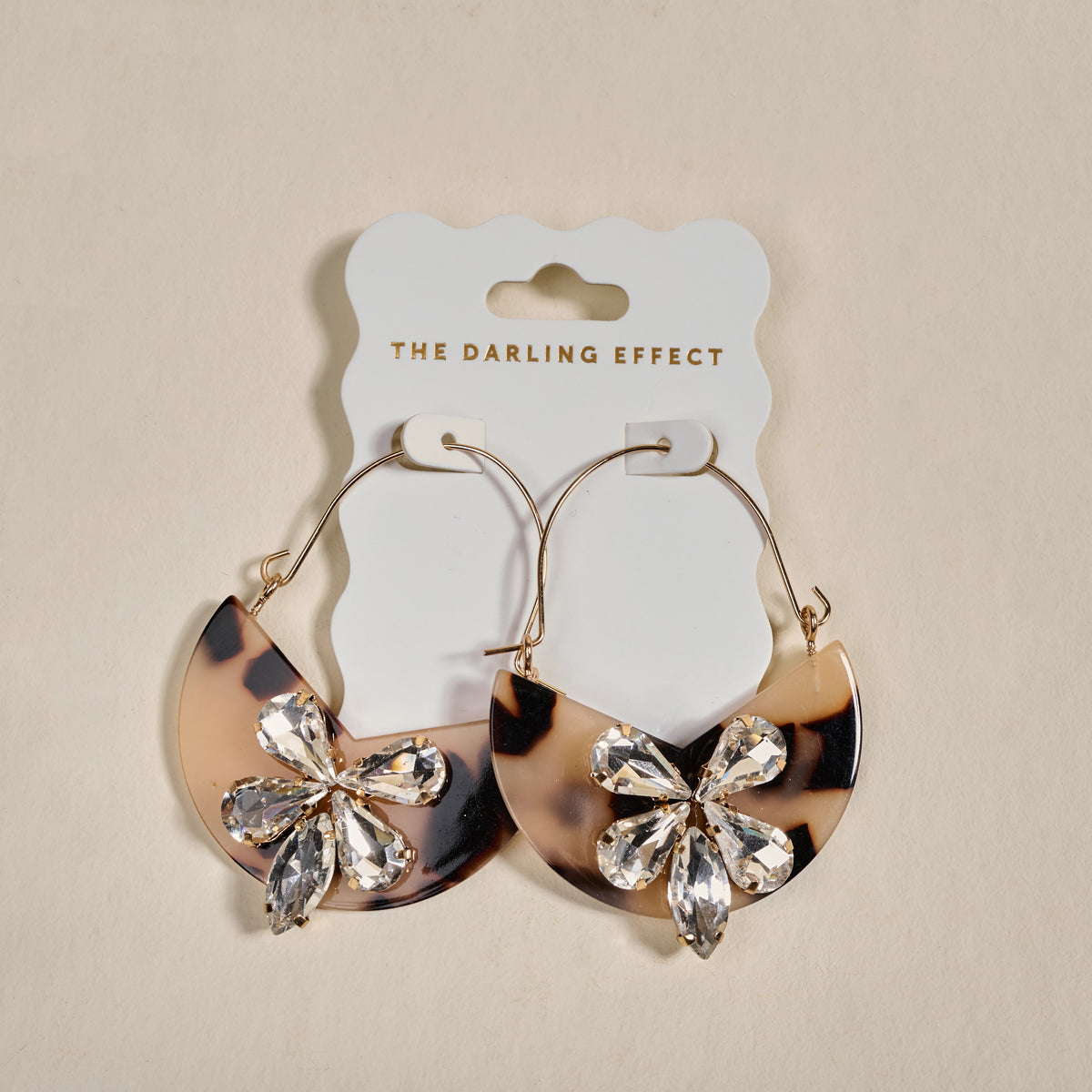 The Darling Effect’s Blonde Tortoise Marble Bloom Hoop Earrings are lightweight gold hoops with half-circle tortoiseshell pendants and crystal flower accents, displayed on a white “The Darling Effect” card.
