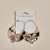 The Darling Effect’s Blonde Tortoise Marble Bloom Hoop Earrings are lightweight gold hoops with half-circle tortoiseshell pendants and crystal flower accents, displayed on a white “The Darling Effect” card.