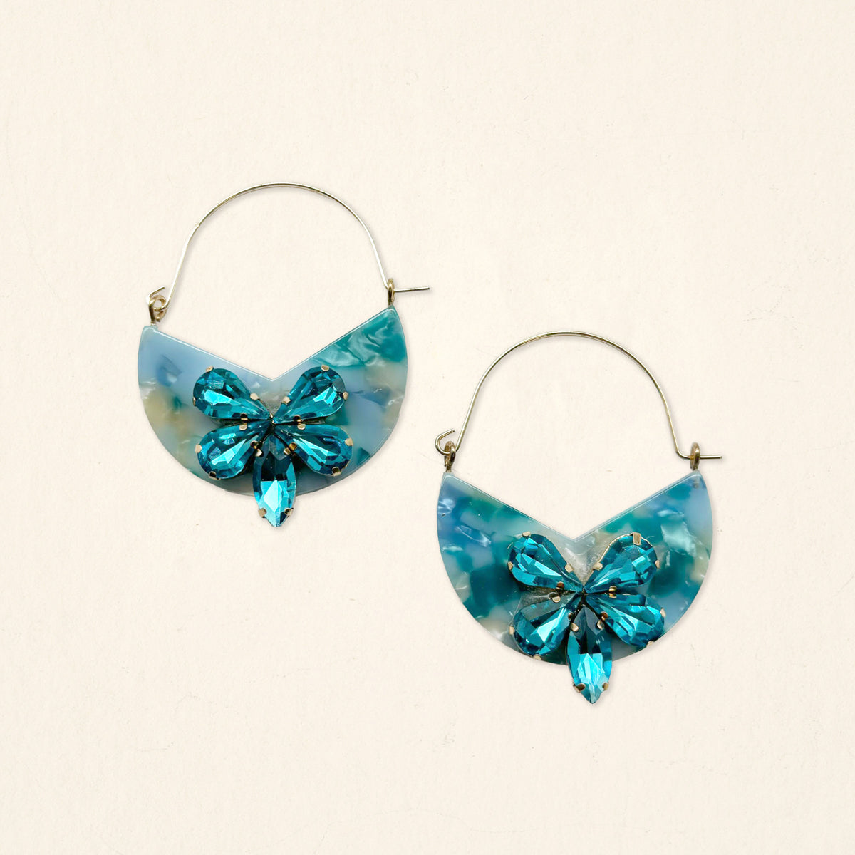 The Darling Effect’s Sea Glass Marble Bloom Hoop Earrings are lightweight statement pieces with glossy acetate hoops, blue marbled semi-circular pendants, and a central blue crystal butterfly, shown on a plain light background.