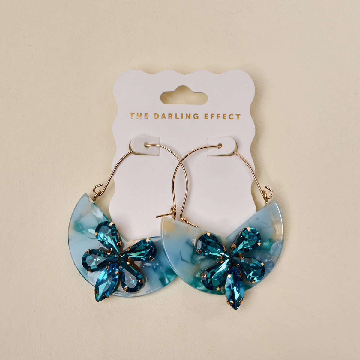 The Darling Effect’s Sea Glass Marble Bloom Hoop Earrings feature lightweight semi-circular blue marbled acrylic pendants and blue gemstone flowers, displayed on a white THE DARLING EFFECT card.