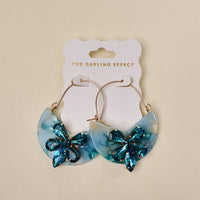 The Darling Effect’s Sea Glass Marble Bloom Hoop Earrings feature lightweight semi-circular blue marbled acrylic pendants and blue gemstone flowers, displayed on a white THE DARLING EFFECT card.