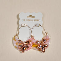 The Darling Effect Sunset Kiss Marble Bloom Hoop Earrings feature crescent-shaped pink, gold, and white acrylic with large pink rhinestone flower accents. Lightweight statement earrings displayed on a branded card, perfect for adding flair to any outfit.