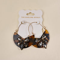 The Darling Effect’s Totally Tortoise Marble Bloom Hoop Earrings feature large gold hoops with brown tortoiseshell crescents and rhinestone butterfly accents—stylish, modern statement pieces on a white branded card.