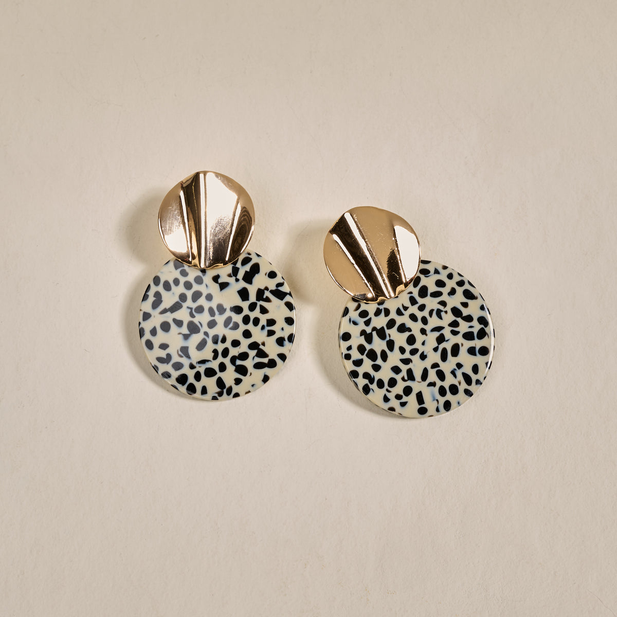 The Darling Effect’s Dalmatian Darling Marble Muse Earrings feature shiny gold-toned tops and bold round black-and-white dalmatian print dangles, shown against a plain beige background.