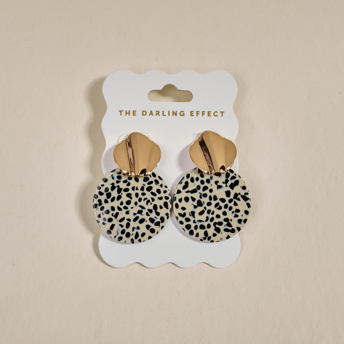 Dalmatian Darling Marble Muse Earrings by The Darling Effect feature gold-tone, lightweight statement circles with a dalmatian print, elegantly displayed on a white scalloped card branded THE DARLING EFFECT.
