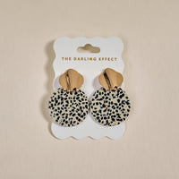 Dalmatian Darling Marble Muse Earrings by The Darling Effect feature gold-tone, lightweight statement circles with a dalmatian print, elegantly displayed on a white scalloped card branded THE DARLING EFFECT.