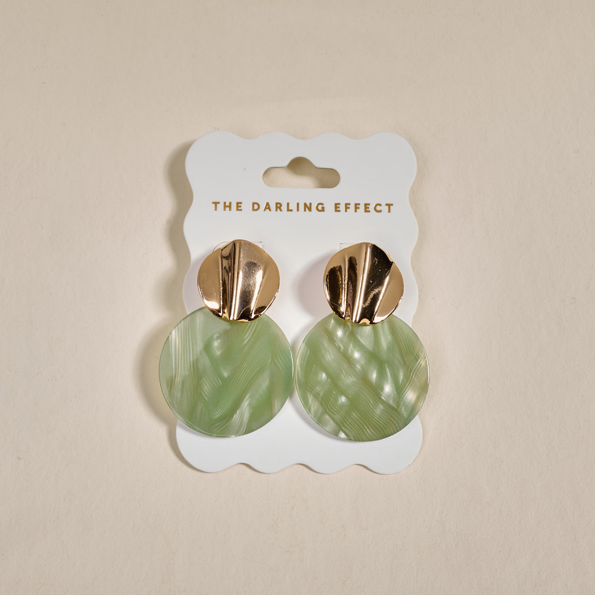 The Darling Effect’s Sage Serenity Marble Muse Earrings feature gold-tone tops and large, round, light green marbled acrylic dangles, displayed on a branded white card.