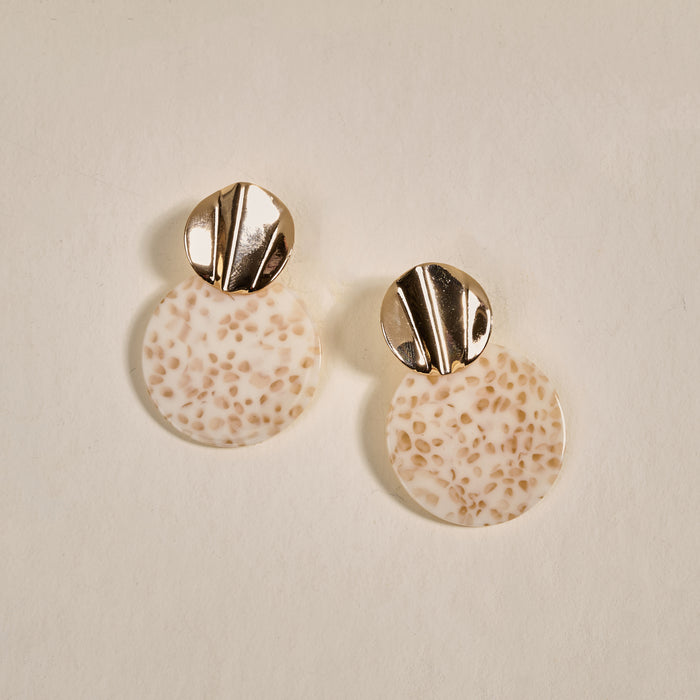 The Darling Effect's Toffee Glow Marble Muse Earrings feature round, textured beige resin discs with speckled patterns and gold-toned, grooved studs, displayed on a light, neutral background.