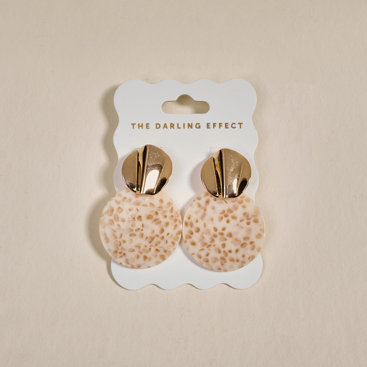 The Darling Effect's Toffee Glow Marble Muse Earrings feature gold metallic tops and round, cream-toned textured dangles, shown on a branded card against a neutral background.