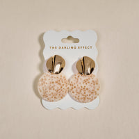 The Darling Effect's Toffee Glow Marble Muse Earrings feature gold metallic tops and round, cream-toned textured dangles, shown on a branded card against a neutral background.