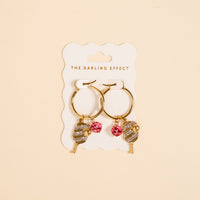 The Darling Effect’s Dinking Around Signature Hoop Earrings feature gold hoops with pickleball paddle and pink gemstone charms, adding playful sparkle to your everyday style.