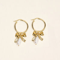 The Darling Effect’s Sweetheart Pearl Signature Hoop Earrings: lightweight gold hoops adorned with delicate bow charms and white teardrop-shaped beads, shown on a light beige background.