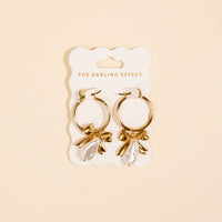 The Darling Effect Sweetheart Pearl Signature Hoop Earrings are lightweight gold hoops with bow accents and teardrop pearls, shown on a white card labeled The Darling Effect against a beige background.