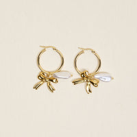 The Darling Effect's Sweetheart Pearl Signature Hoop Earrings are lightweight gold hoops with decorative gold bows and white teardrop-shaped pearls, elegantly displayed on a plain light background.