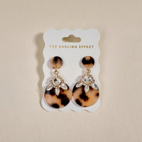 The Darling Effect's Blonde Tortoise Show Stopper Earrings feature rhinestone-embellished acetate in a statement design, displayed on a white THE DARLING EFFECT card with gold text against a beige background.