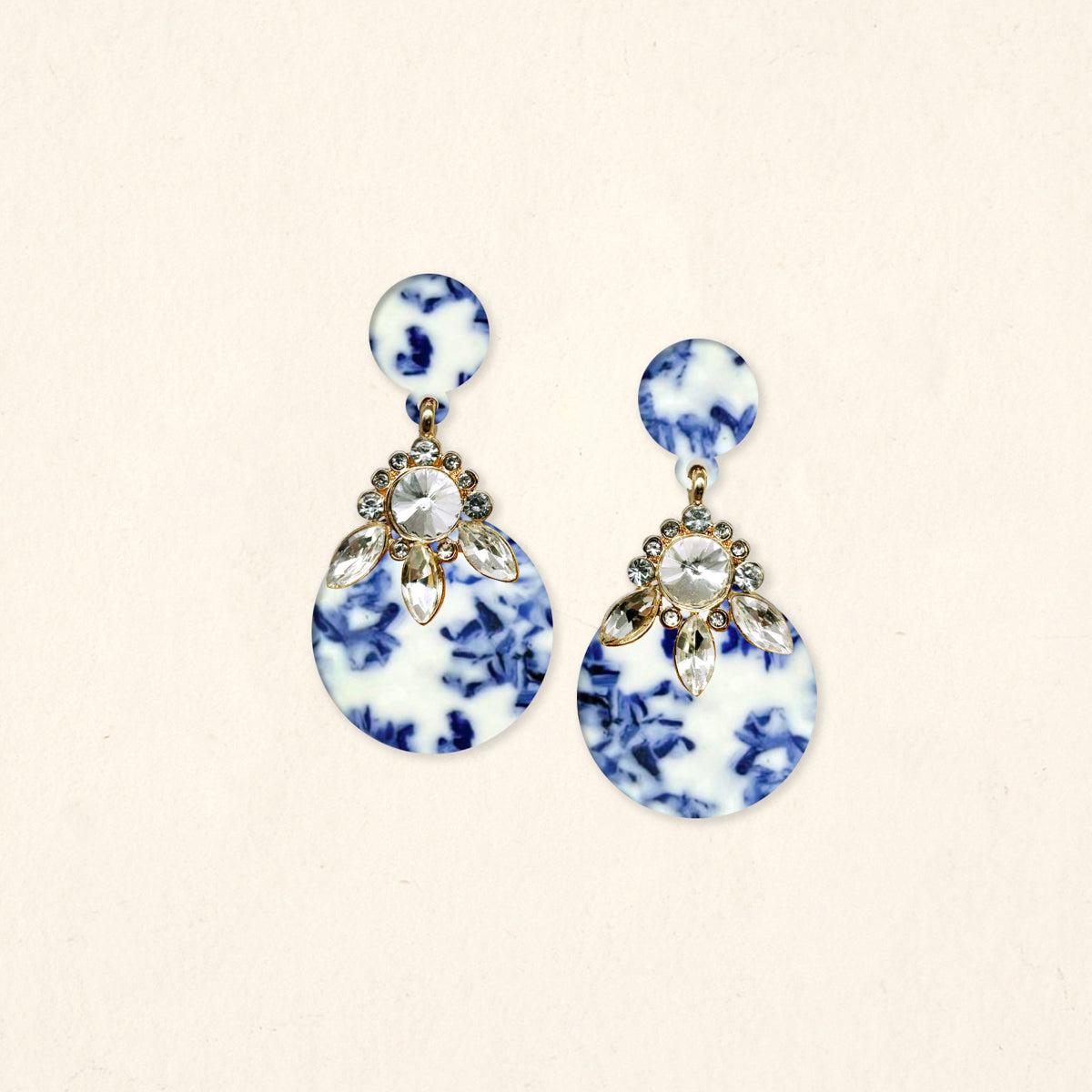 The Darling Effect’s Chinoiserie Flair Show Stopper Earrings feature round marbled blue and white acetate with clear teardrop crystals atop the larger circle—lightweight statement earrings perfect for standout style.