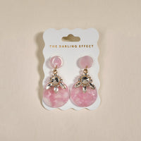 The Rosé Radiance Show Stopper Earrings by The Darling Effect are pink, lightweight statement earrings with rhinestone accents, displayed on a white card with gold branding against a neutral beige backdrop.