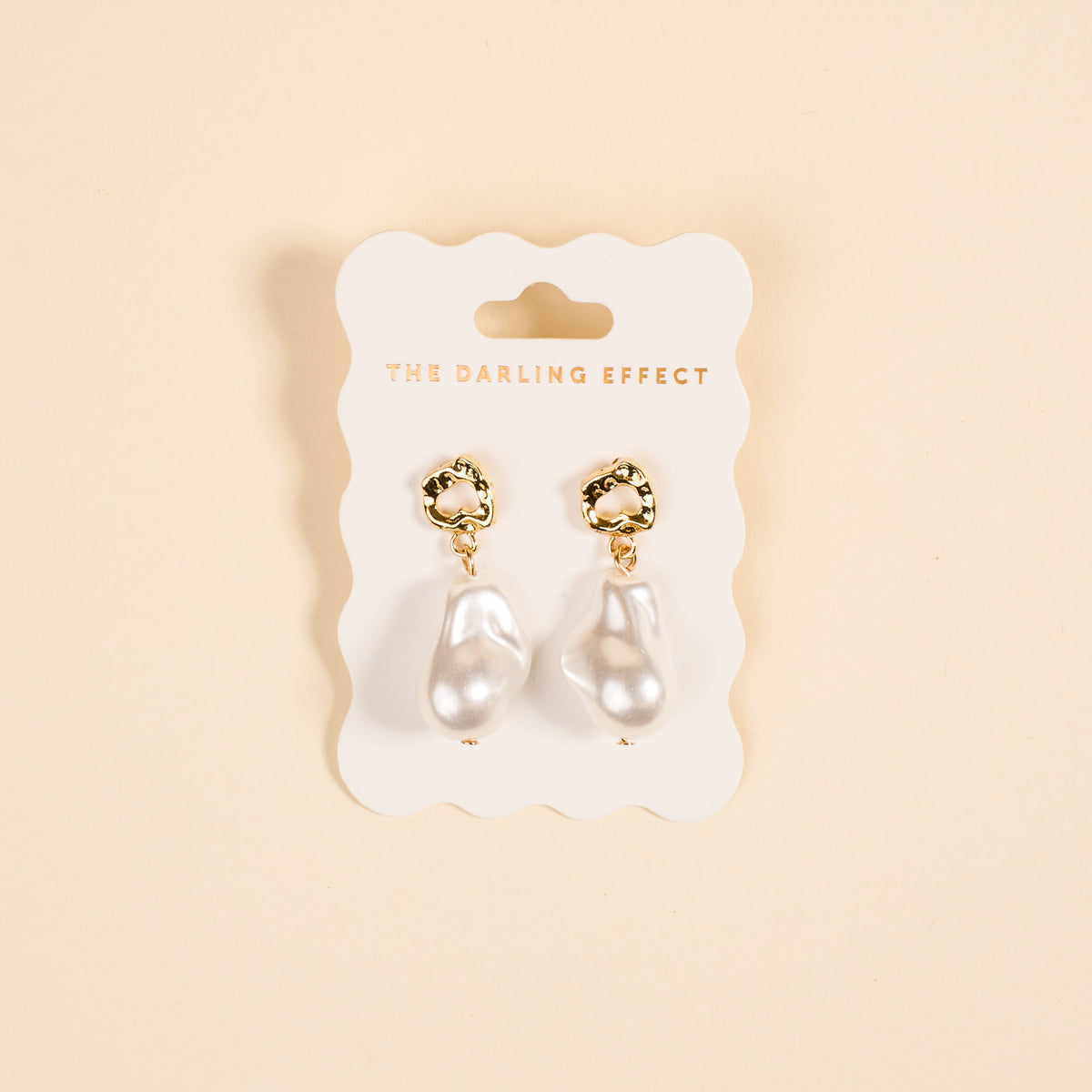 The Darling Effect Baroque Pearl Standout Earrings, featuring high-shine gold and irregular white pearl drops, are displayed on a branded white card against a light beige background.