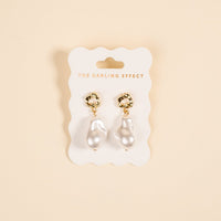 The Darling Effect Baroque Pearl Standout Earrings, featuring high-shine gold and irregular white pearl drops, are displayed on a branded white card against a light beige background.