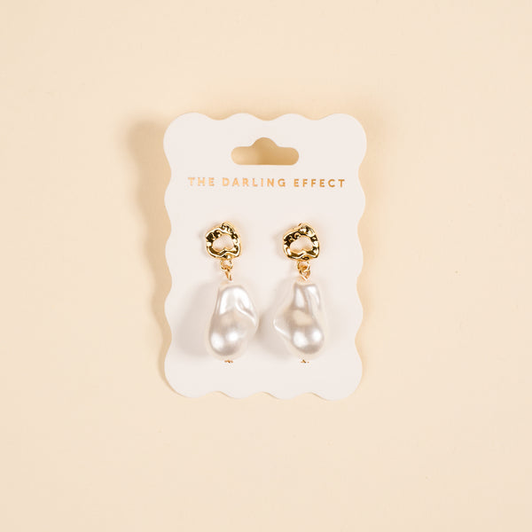 The Darling Effect Baroque Pearl Standout Earrings, featuring high-shine gold and irregular white pearl drops, are displayed on a branded white card against a light beige background.