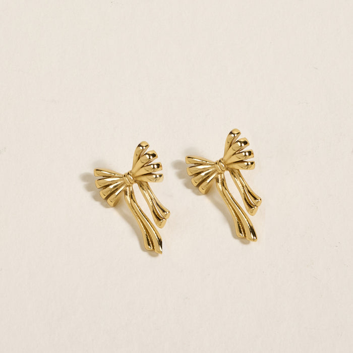 The Darling Effect’s Stay Classy Bow Standout Earrings feature golden, bow-shaped designs with flowing ribbon ends and a high-shine finish, shown on a light beige background.