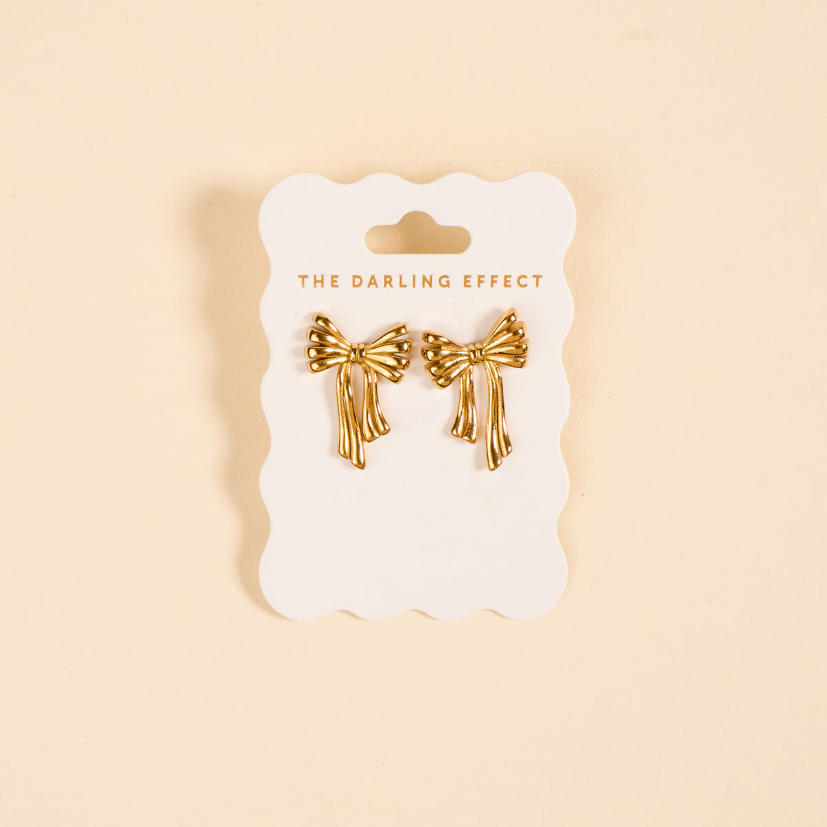 The Darling Effect Stay Classy Bow Standout Earrings, featuring a high-shine gold bow design, are displayed on a branded white card against a beige background.