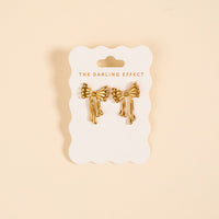 The Darling Effect Stay Classy Bow Standout Earrings, featuring a high-shine gold bow design, are displayed on a branded white card against a beige background.