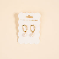 The Dainty Drift Standout Earrings by The Darling Effect feature gold-tone shine and dangling white pearls, displayed on a branded card against a beige background—ideal for adding elegance to any special occasion.