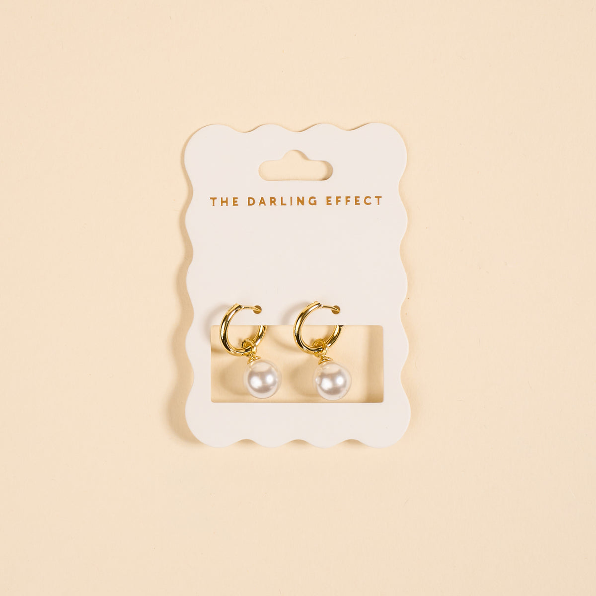 The Darling Effect’s Dainty Pearl Standout Earrings feature golden hoops with pearl drops, elegantly displayed on a scalloped white card—perfect for making a statement.