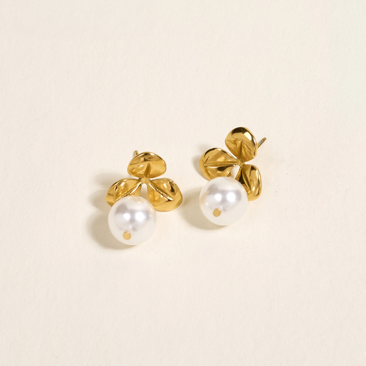 The Darling Effect's Fleur de Pearl Standout Earrings feature three gold petal shapes above a round white pearl with a high-shine finish, showcased on a light cream background.