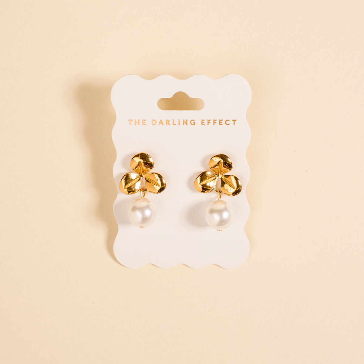 The Fleur de Pearl Standout Earrings by The Darling Effect feature a floral design with dangling white pearls and a high-shine finish, displayed on a branded white card against a beige background.