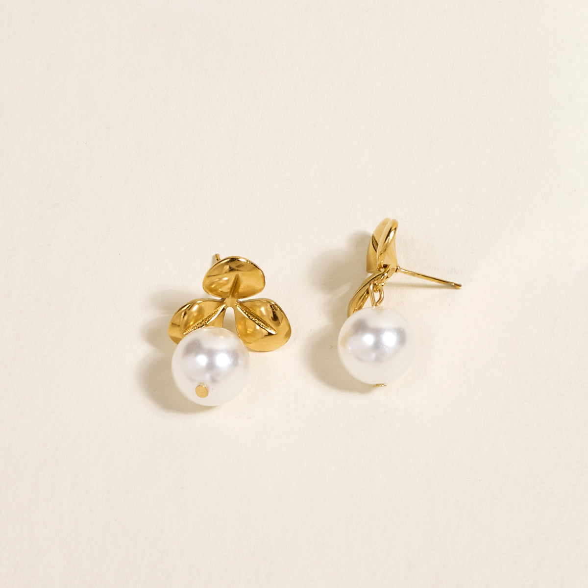 The Darling Effect's Fleur de Pearl Standout Earrings feature three golden leaves and a round white pearl on each, showcased on a light cream background—an elegant statement accessory for any occasion.
