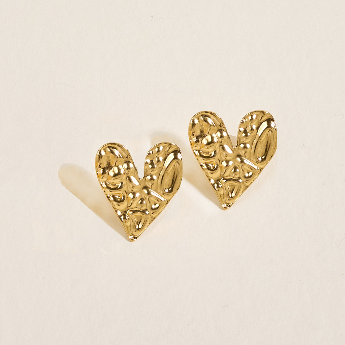 The Darling Effect’s Hammered Heart Standout Earrings—gold-toned and textured in a heart shape—are shown on a light, neutral background with soft left shadows, perfect for making a statement with high-shine jewelry.