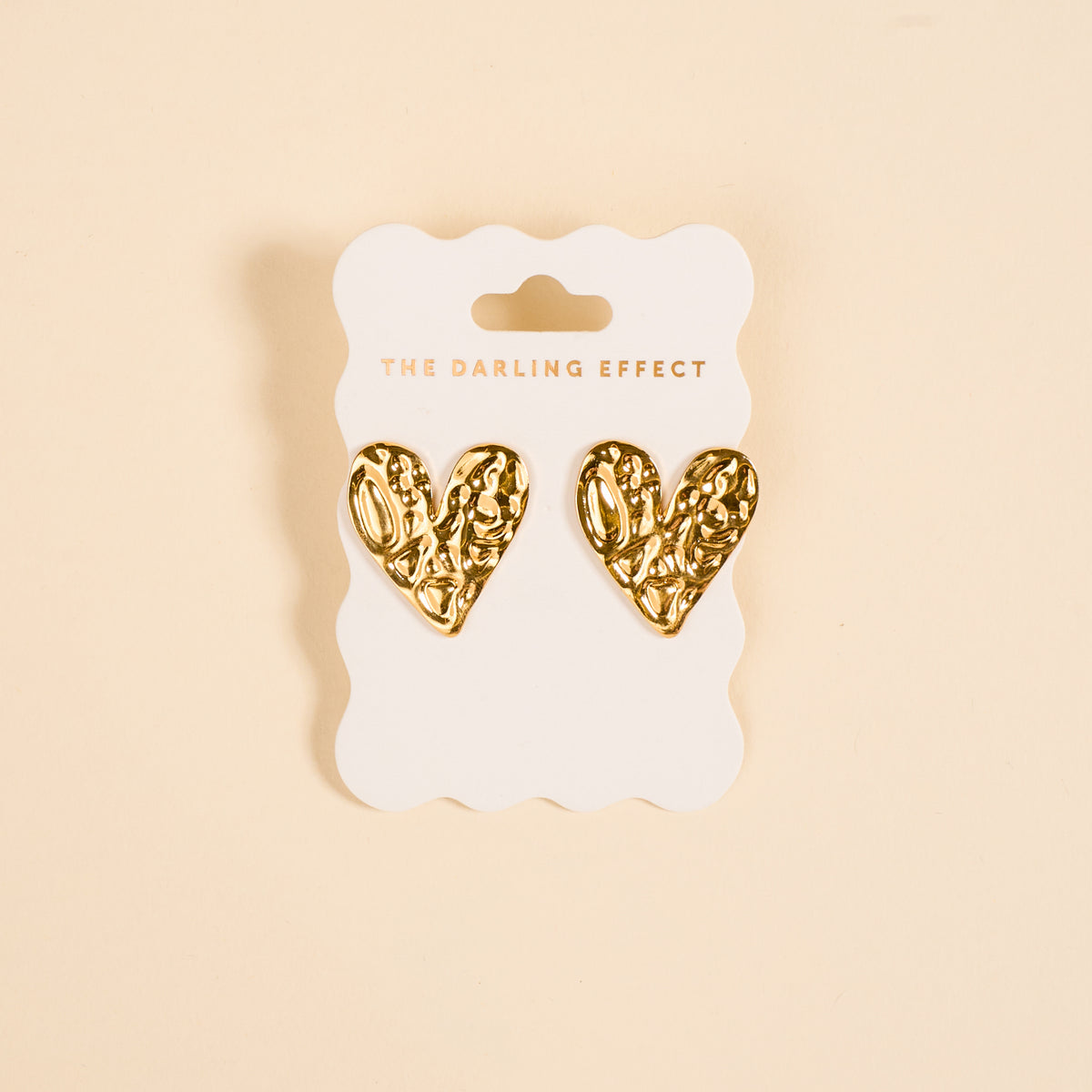 The Darling Effect's Hammered Heart Standout Earrings feature textured gold, heart-shaped designs with a high-shine finish, displayed on a scalloped white card—ideal for anyone who loves statement jewelry.