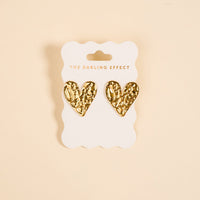 The Darling Effect's Hammered Heart Standout Earrings feature textured gold, heart-shaped designs with a high-shine finish, displayed on a scalloped white card—ideal for anyone who loves statement jewelry.