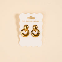 The Heirloom Standout Earrings by The Darling Effect, featuring a teardrop-shaped top and high-shine finish, are displayed on a white scalloped card with the brand name against a beige background.