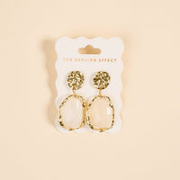 The Darling Effect's Liquid Gold Standout Earrings feature textured gold tops, irregular oval-shaped white stones, and a high-shine finish. Displayed on a branded card.