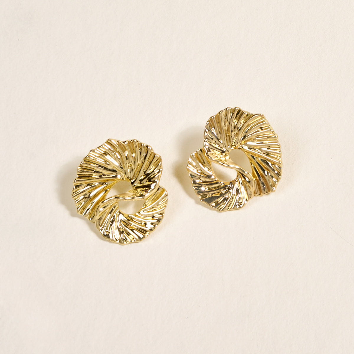 The Darling Effect's Ripple Effect Standout Earrings are golden, circular statement pieces with a textured, twisted design, shown on a light surface.