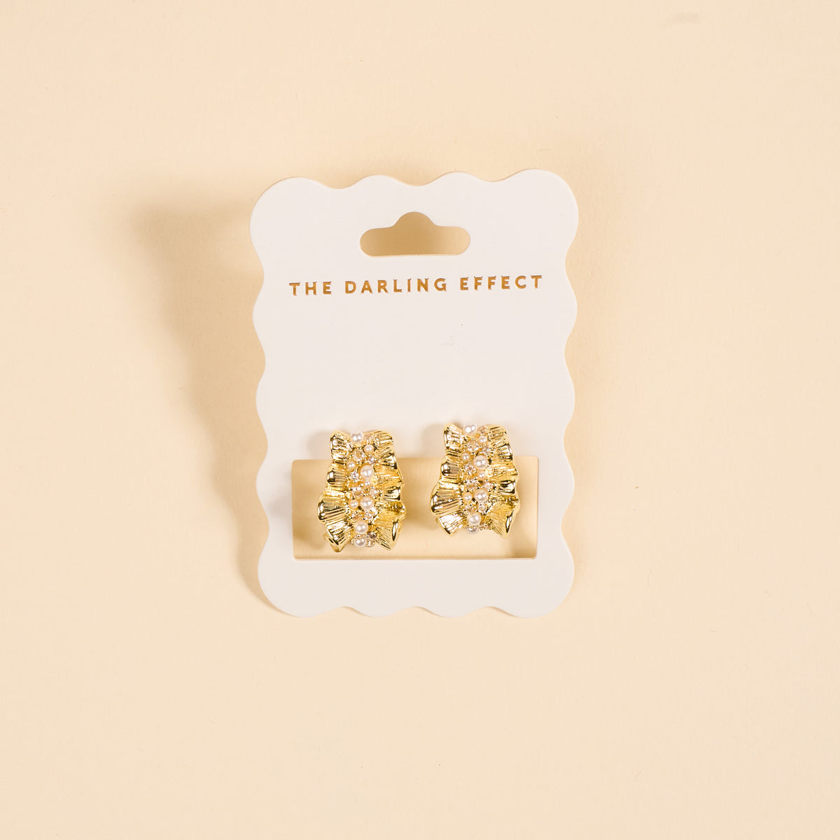Golden Ruffle Standout Earrings by The Darling Effect feature textured gold studs with pearl and crystal accents, displayed on a white scalloped card against a soft peach background.