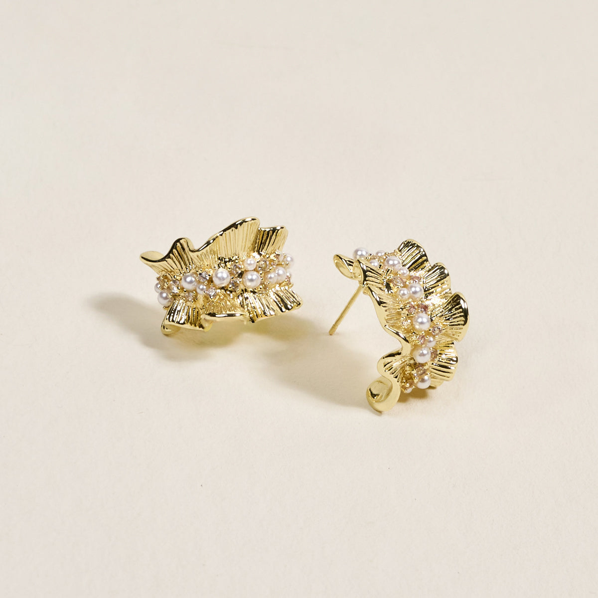 The Darling Effect's Golden Ruffle Standout Earrings feature a high-shine finish and pearl accents along the center, displayed on a light beige background.