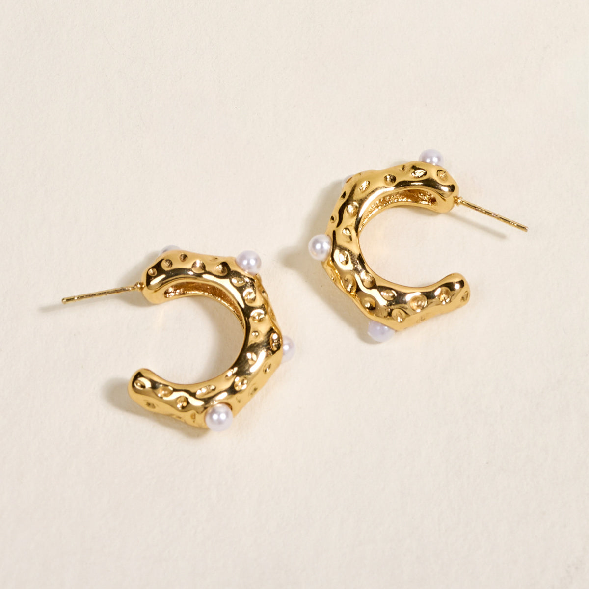 The Darling Effect's Say It All Standout Earrings feature textured golden hexagonal hoops with small white pearls and a high-shine finish, beautifully displayed on a cream-colored surface.