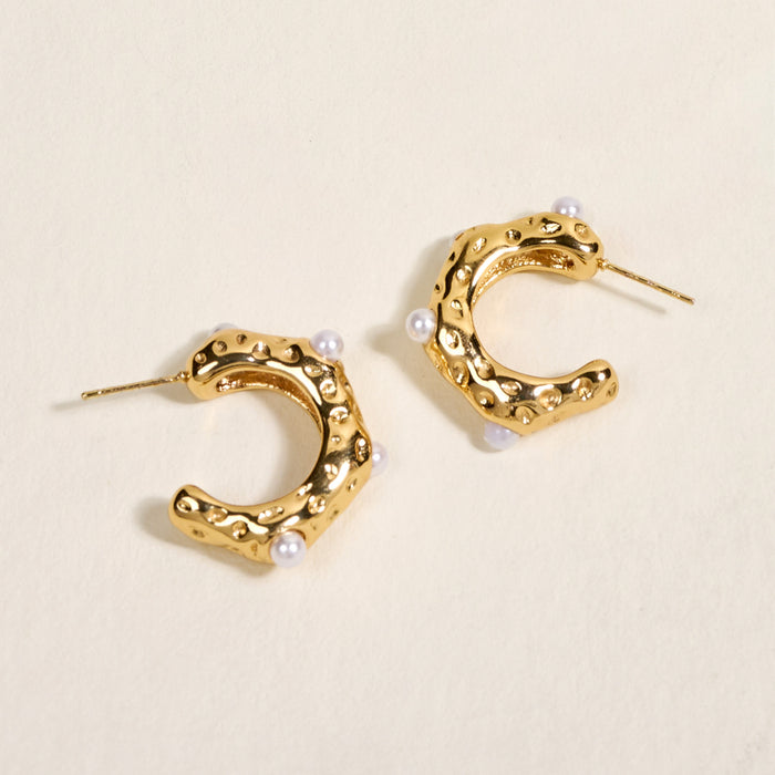 The Darling Effect's Say It All Standout Earrings feature textured golden hexagonal hoops with small white pearls and a high-shine finish, beautifully displayed on a cream-colored surface.