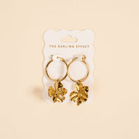 Sunlit Petal Standout Earrings by The Darling Effect feature high-shine golden finish and dangling flower charms, displayed on a white card—perfect for special occasions against a soft beige background.