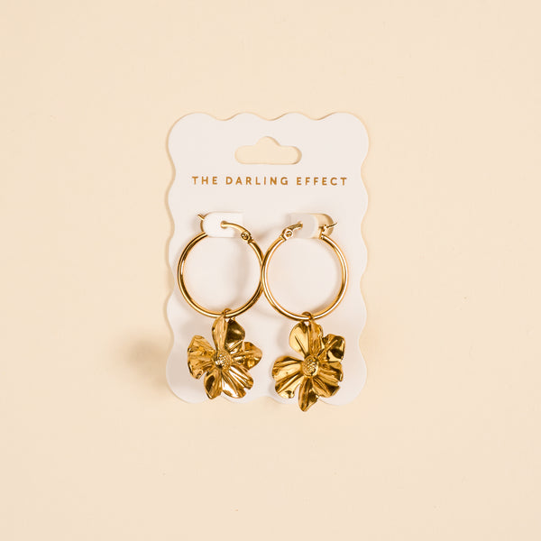 Sunlit Petal Standout Earrings by The Darling Effect feature high-shine golden finish and dangling flower charms, displayed on a white card—perfect for special occasions against a soft beige background.