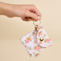 A hand holds the-darling-effect’s Endless Daydream Cream Puffy Earbud Case Keychain, featuring a pink and orange floral pattern, gold button snap, gold keyring, clasp for easy carrying, and designed to fit white wireless earbuds.