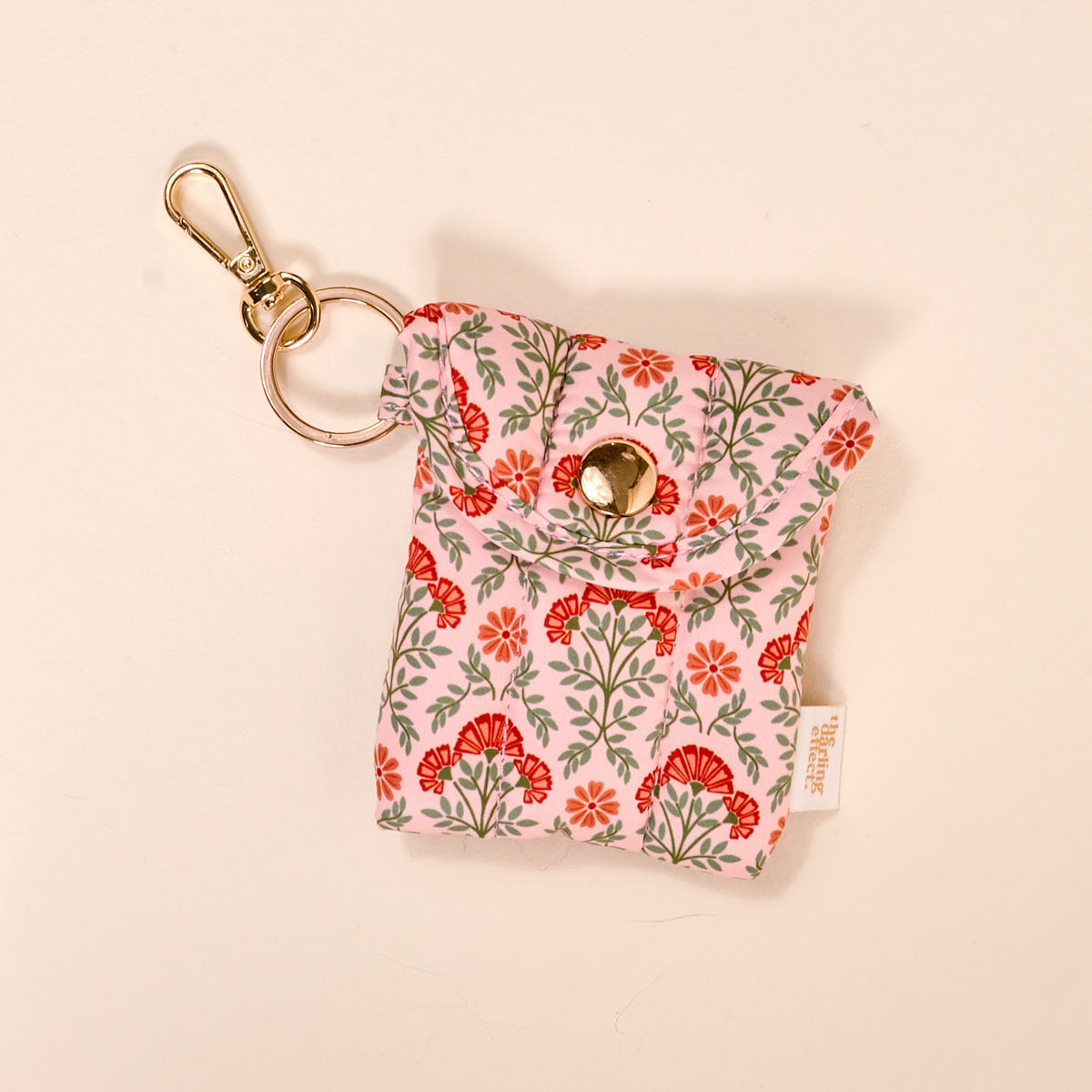 The Darling Effect Everbloom Dusty Rose Puffy Earbud Case features a red and green floral pattern, snap closure, and gold keyring clip. Shown against a light beige background, it's perfect as an earbud keychain.