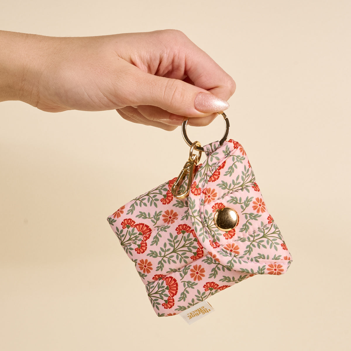 A hand holds The Darling Effect Everbloom Dusty Rose Puffy Earbud Case, featuring a floral quilted design, gold key ring, and snap button closure, against a beige background.