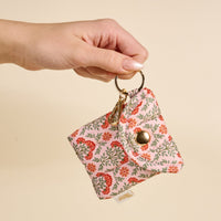 A hand holds The Darling Effect Everbloom Dusty Rose Puffy Earbud Case, featuring a floral quilted design, gold key ring, and snap button closure, against a beige background.